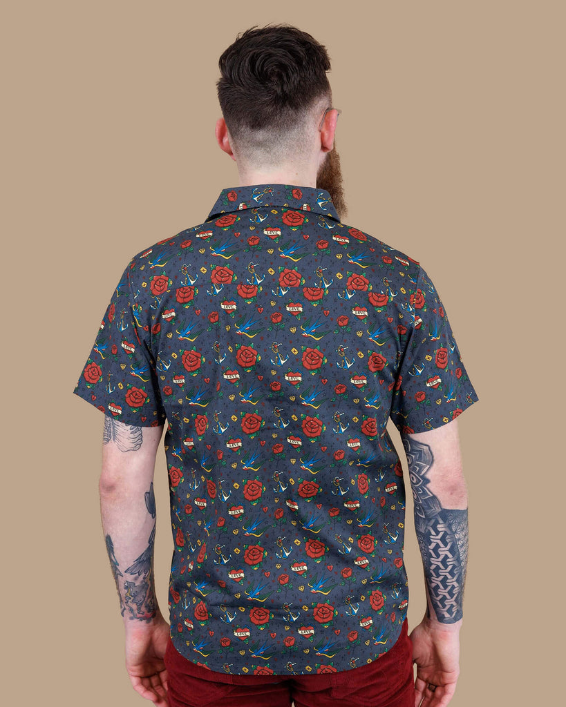 Dec, a white male model with a beard and glasses is wearing Retro Old School Tattoo Print Short Sleeve Shirt with Tawney Port Corduroy Bell Bottom Flares. The print has traditional tattoo style red roses, blue swallows, yellow envelopes, anchors and valentines love heart. Dec is stood in front of a beige backdrop at a photography studio in Hove with his back to the camera and his arms down by his sides.
