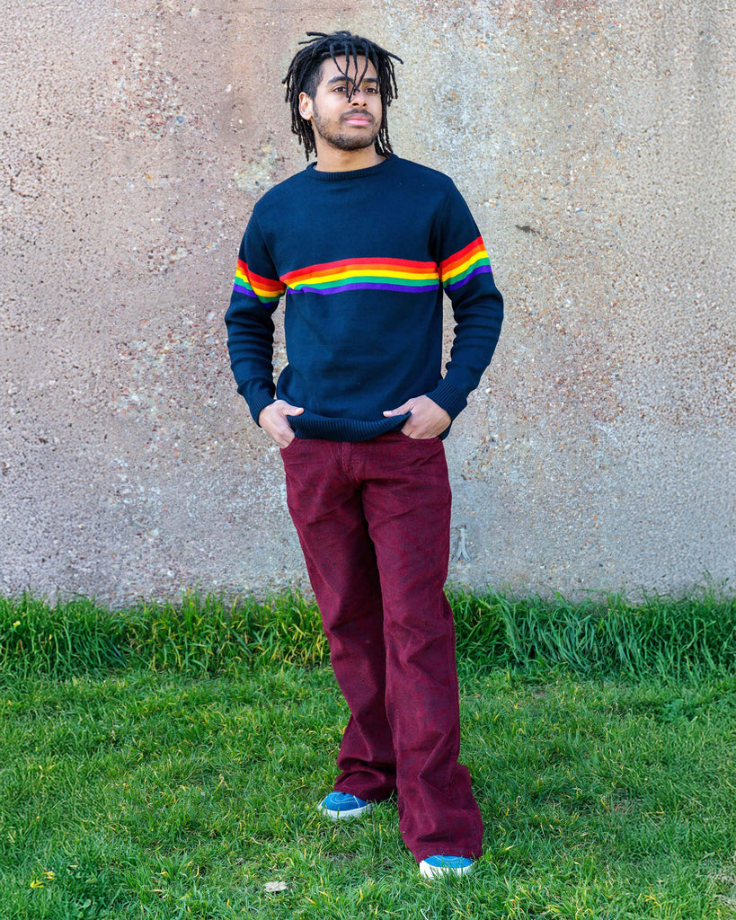 Basil is stood in front of a plain brick wall wearing the tawney port paisley corduroy bell bottom super flares with the navy retro rainbow stripe jumper and blue trainers. He is facing forward with both hands resting in the front pockets whilst looking off to the right.