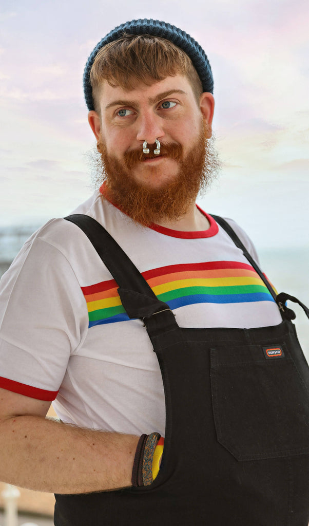 Model is stood near a beach wearing the white Riner retro rainbow striped tshirt with black dungarees and a blue beanie. They are facing to the right looking over their shoulder to the left smiling with both hands inside the dungarees. Photo is cropped from the waist up.