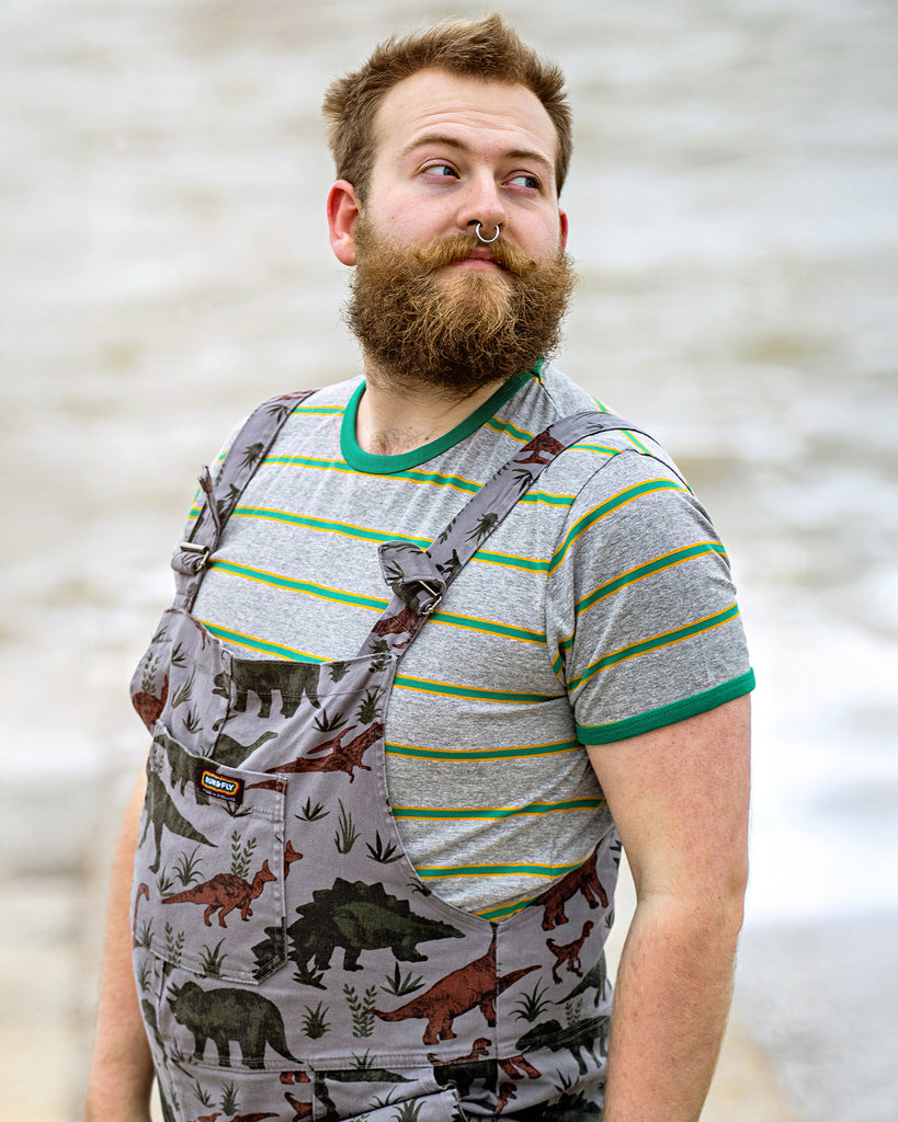 Thomas is stood on the seafront wearing the retro mod ash grey engineered striped tshirt with the adventure dinosaur dungarees. They have a brown beard and septum ring. They're facing to the left posing with both arms resting by their side and looking over their shoulder to the right. Photo is cropped from the hips up.