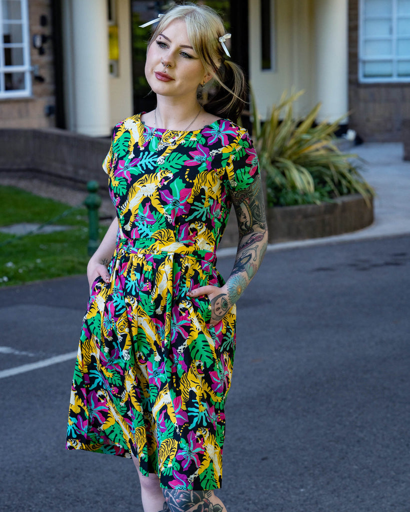 Tattooed model wearing a Run & Fly tiger print tea dress with a fitted waist and pockets. The bold print includes yellow tigers, pink flowers and green leaves. Ethical fashion from UK brand Run & Fly.