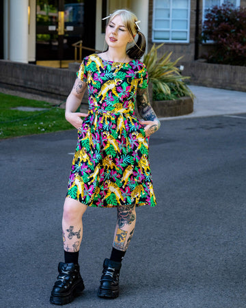 Model with tattoos and pigtails wearing a colourful Run & Fly tiger print tea dress. The short sleeve dress features pink flowers, green leaves and yellow tigers. A playful ethical piece from UK brand Run & Fly.