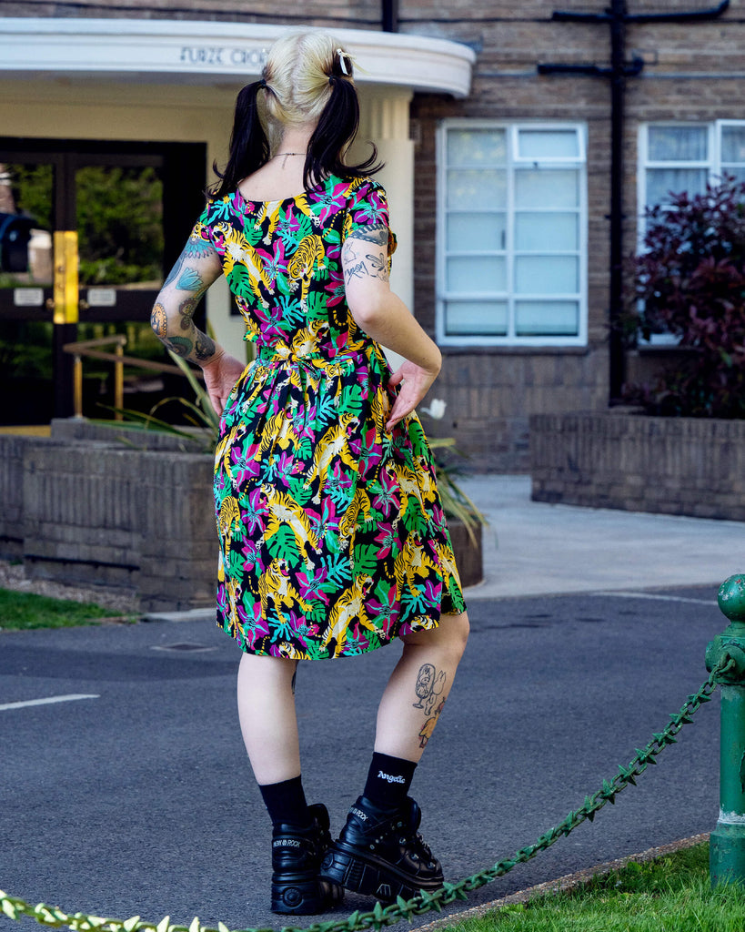 Model with black and blonde pigtails wearing a Run & Fly tiger print tea dress, seen from the back. The dress features vibrant yellow tigers, pink flowers and green monstera leaves on a black background. Ethically made by UK slow fashion brand Run & Fly.