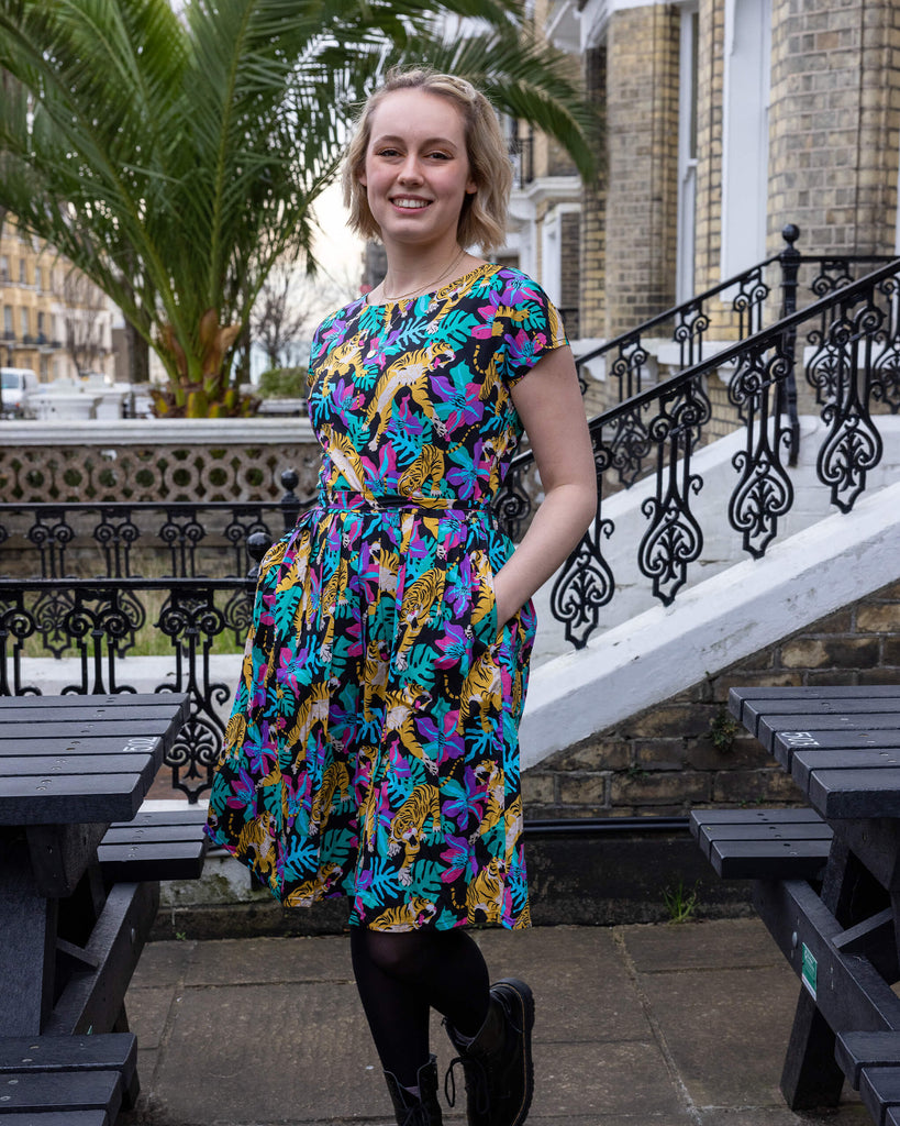 Amy is wearing the black tiger lily stretch belted tea dress, in teal blue orange pink and purple, below the knee with round neck. Model is looking at the camera and smiling with hands in their pockets. Black ornate railing in background with black bench and big plant palm tree in background.