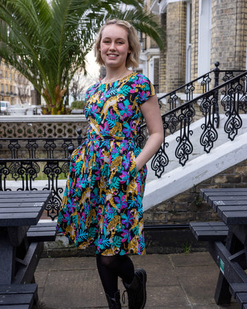 Amy is wearing the black tiger lily stretch belted tea dress, in teal blue orange pink and purple, below the knee with round neck. Model is looking at the camera and smiling with hands in their pockets. Black ornate railing in background with black bench and big plant palm tree in background.