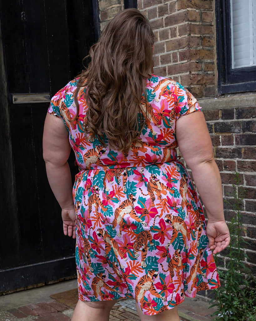 Natalie is wearing Tiger Lily Stretch Belted Tea Dress with Pockets with cactus shaped earrings and pink boots. The baby pink dress has an all over print of tigers with flowers and leaves. Natalie is stood outside in Hove with her back to the camera and arms downs by her sides.