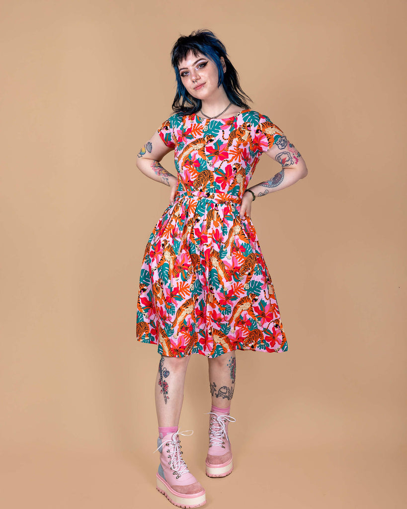 Faeryn is stood in a photography studio in Hove with a beige backdrop wearing Tiger Lily Stretch Belted Tea Dress with PocketsThe baby pink dress has an all over print of tigers with flowers and leaves. They are posing toward the camera with their hands on their hips.