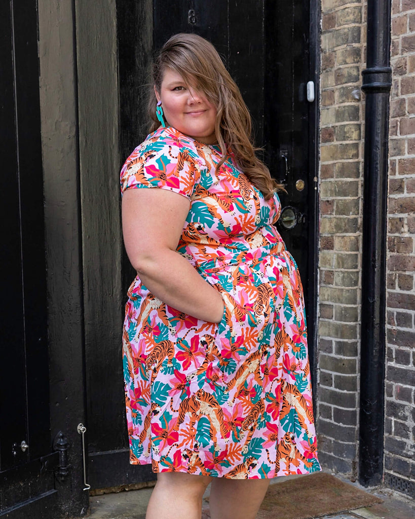 Natalie is wearing Tiger Lily Stretch Belted Tea Dress with Pockets with cactus shaped earrings and pink boots. The baby pink dress has an all over print of tigers with flowers and leaves. Natalie is standing sideways outside in Hove facing the camera with her hands in the pockets of the dress.