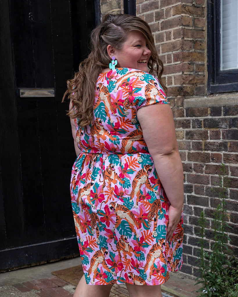 Natalie is wearing Tiger Lily Stretch Belted Tea Dress with Pockets with cactus shaped earrings and pink boots. The baby pink dress has an all over print of tigers with flowers and leaves. Natalie is stood outside in Hove with her back to the camera and is looking back over her shoulder smiling.
