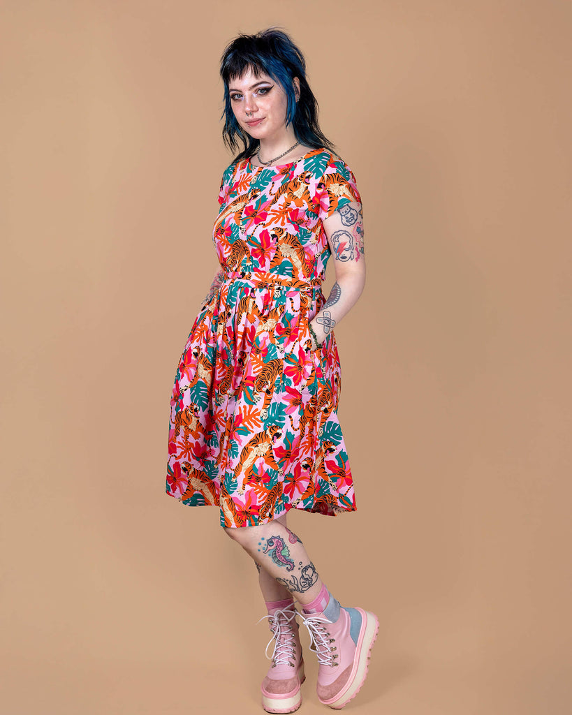 Faeryn is stood in a photography studio in Hove with a beige backdrop wearing Tiger Lily Stretch Belted Tea Dress with Pockets. The baby pink dress has an all over print of tigers with flowers and leaves. Faeryn is posing with one leg slightly bent and is looking at the camera with their hands in the dress pockets.
