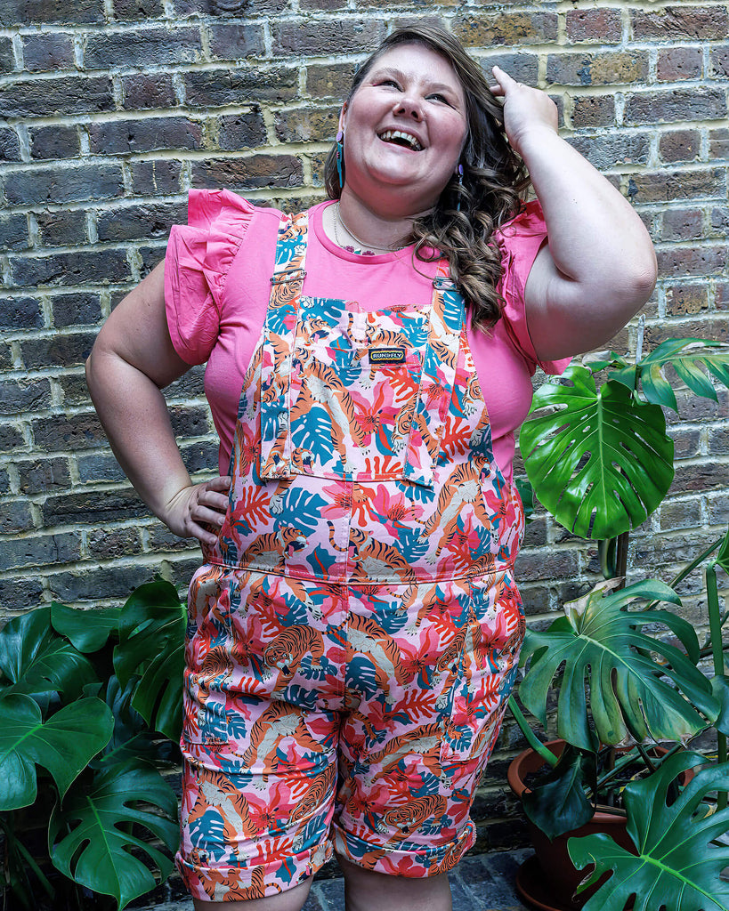 A femme model wearing the tiger lily stretch twill dungaree shorts outside amongst plants. They are smiling and posing towards the camera.