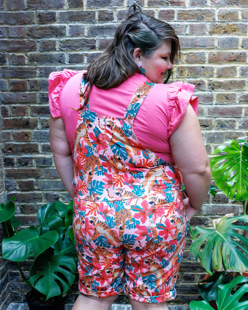 A femme model wearing the tiger lily stretch twill dungaree shorts outside amongst plants. They are smiling and posing towards the camera.