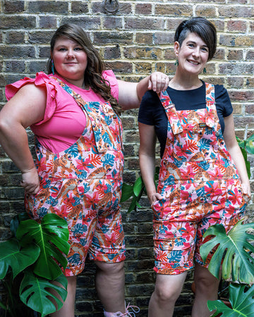 Two femme models wearing the tiger lily stretch twill dungaree shorts outside amongst plants. They are both smiling and posing towards the camera.