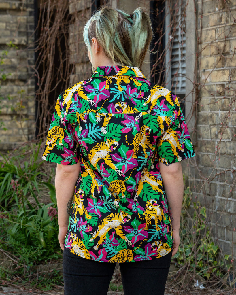 Person with pastel green hair in pigtails wearing a Run & Fly tiger print shirt, standing with back to camera. The shirt features bold yellow tigers, green monstera leaves and pink flowers on a black background. Ethical and vibrant slow fashion by UK brand Run & Fly.
