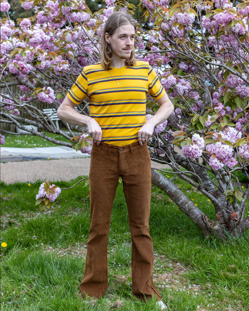  Model Jack stands in front of a blooming cherry blossom tree, wearing Run & Fly’s Tobacco Corduroy Flares paired with a yellow and black striped T-shirt. The earthy tones of the flares complement the natural surroundings.
