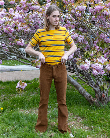  Model Jack stands in front of a blooming cherry blossom tree, wearing Run & Fly’s Tobacco Corduroy Flares paired with a yellow and black striped T-shirt. The earthy tones of the flares complement the natural surroundings.