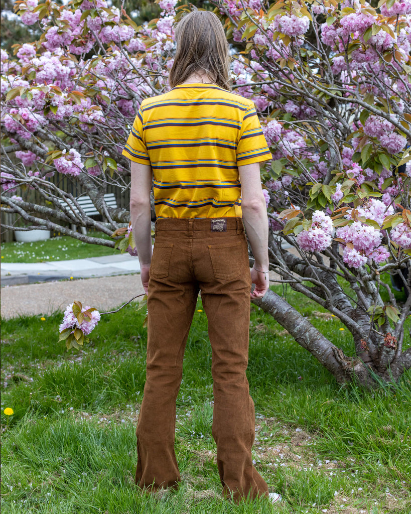  Back view of model Jack wearing Run & Fly’s Tobacco Corduroy Flares, standing in front of a cherry blossom tree. The flares feature a relaxed fit and vintage-inspired style.