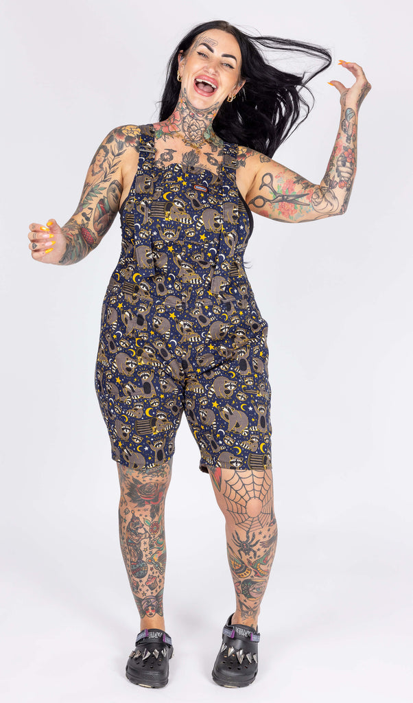 Aimee, a heavily tattooed model with long dark hair is stood in a photography studio in Hove in front of a white backdrop wearing Trash Pandas Stretch Twill Dungaree Shorts with black shoes. The dungaree shorts print features cheeky illustrated racoons, trash bins, rubbish, yellow moons and stars and white sparkles on a dark blue background. Aimee is facing the camera and smiling whilst swishing her hair.