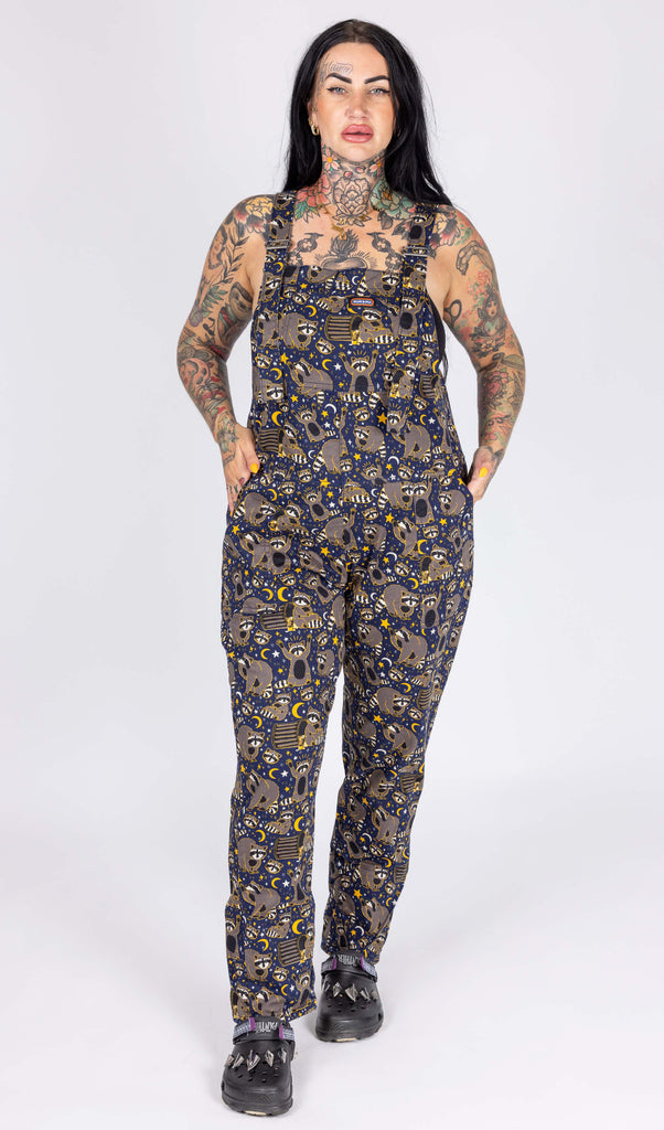 Aimee, a heavily tattooed model with long dark hair is stood in a photography studio in Hove in front of a white backdrop wearing Seconds Trash Pandas Stretch Twill Dungarees with black shoes. The dungaree print features cheeky illustrated racoons, trash bins, rubbish, yellow moons and stars and white sparkles on a dark blue background. Aimee is posing towards the camera with one leg in front of the other and her hands in the pockets of the dungarees. 
