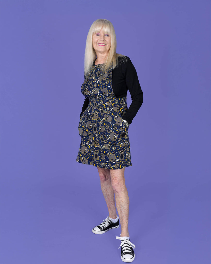 The Trash Pandas Stretch Twill Pinafore Dress worn by a femme model with blonde hair, a black long sleeve tshirt and black converse style trainers on a purple studio background. She is facing forward leaning back on one leg smiling with both hands resting in the side pockets. The print is a rich dark blue with grey racoons, trash bins, white and yellow sparkles, yellow stars and moons and small eyes.