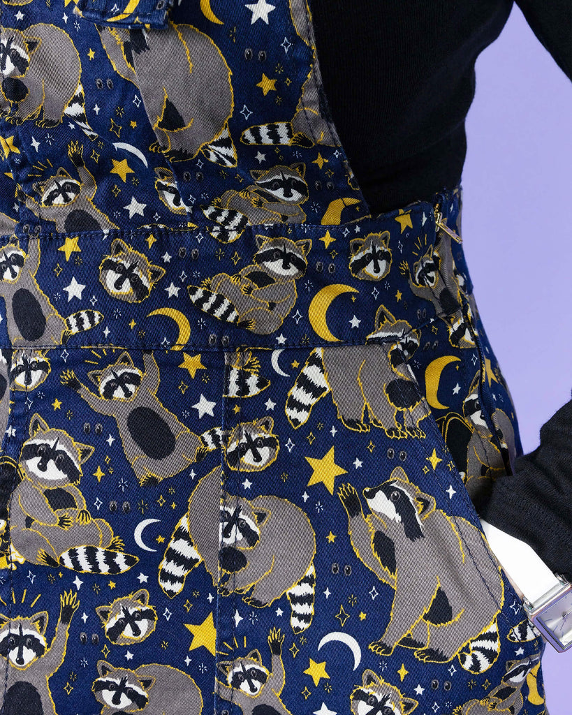 Close up of the Trash Pandas Stretch Twill Pinafore Dress worn by a femme model with a black long sleeve tshirt on a purple studio background. She is facing to the left with her hand resting in the pinafore side pocket. The print is a rich dark blue with grey racoons, trash bins, white and yellow sparkles, yellow stars and moons and small eyes.