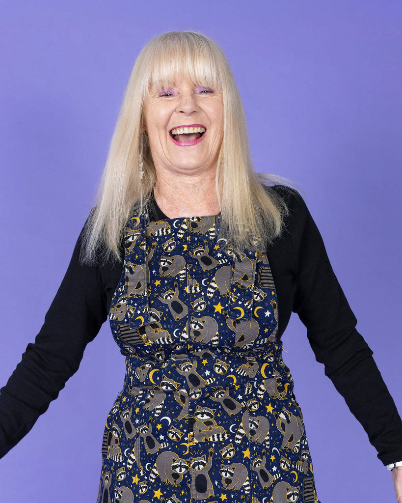 Close up of the Trash Pandas Stretch Twill Pinafore Dress worn by a femme model with blonde hair and a black long sleeve tshirt on a purple studio background. She is facing forward laughing with her hands out to the side. The print is a rich dark blue with grey racoons, trash bins, white and yellow sparkles, yellow stars and moons and small eyes.