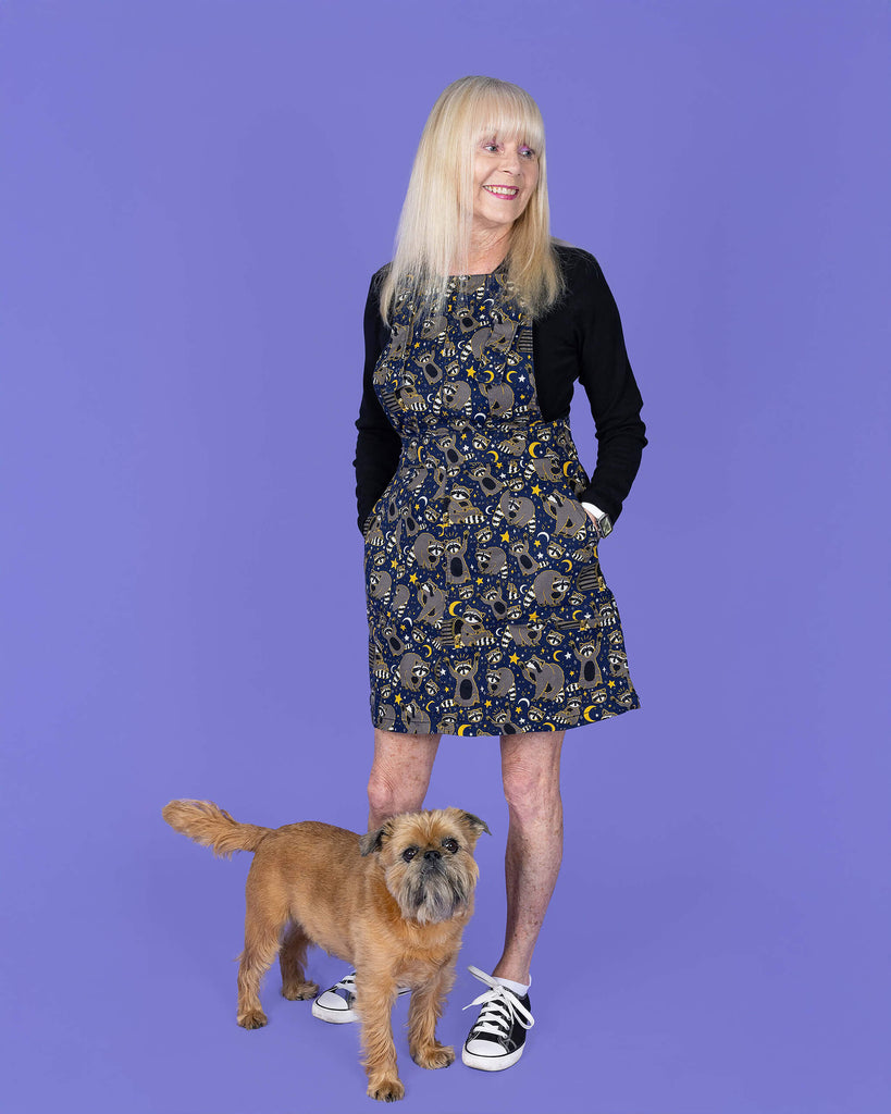 The Trash Pandas Stretch Twill Pinafore Dress worn by a femme model with blonde hair, a black long sleeve tshirt and black converse style trainers on a purple studio background with a small brown dog at her feet. She is facing forward smiling looking off to the right with both hands resting in the side pockets. The print is a rich dark blue with grey racoons, trash bins, white and yellow sparkles, yellow stars and moons and small eyes.