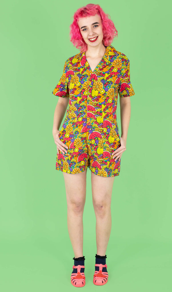 The Tutti Frutti Stretch Twill Playsuit on a femme model with short pink curly hair with pink jelly shoes and black socks. She is stood on a green background facing forward smiling with both hands resting in the pockets. The playsuit print is a blue background full of bright colourful fruit illustrations of limes, lemons, oranges, apples, berries and watermelon.