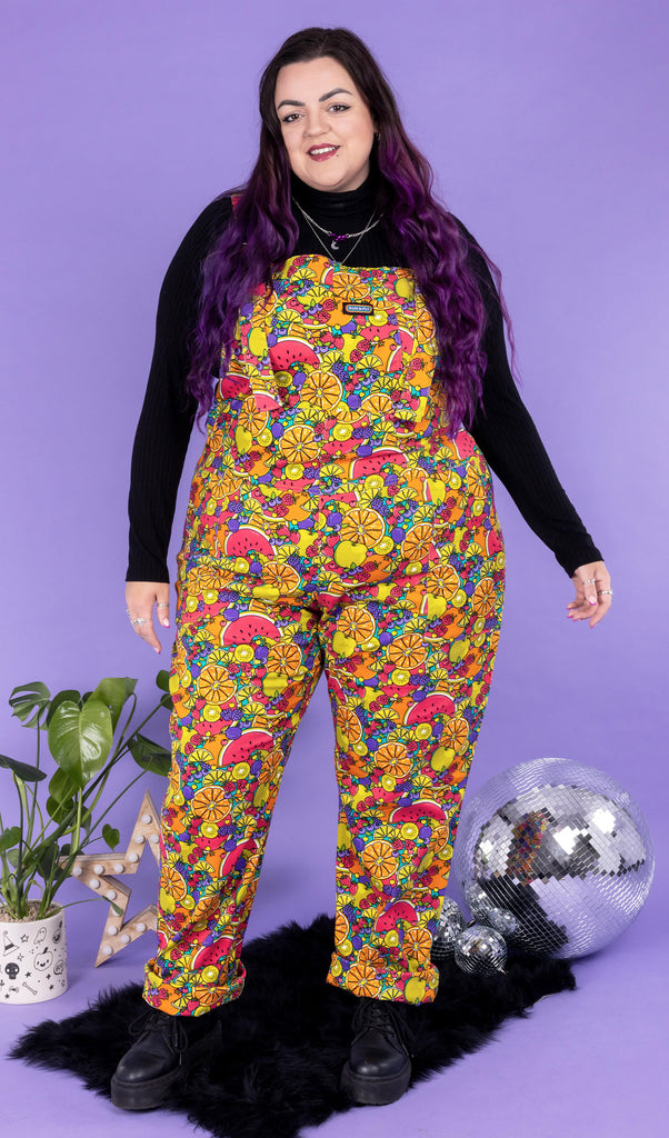Luisa, a white femme model with purple hair, is wearing Tutti Frutti Stretch Twill Dungarees with a long sleeve black turtle neck underneath and black boots. The dungarees have a Jen James design of various fruit all over in bright red, orange, purple, yellow and green. Luisa is stood in front of a lilac background on a black fluffy rug amongst disco balls, a star shaped light and a plant facing the camera smiling.
