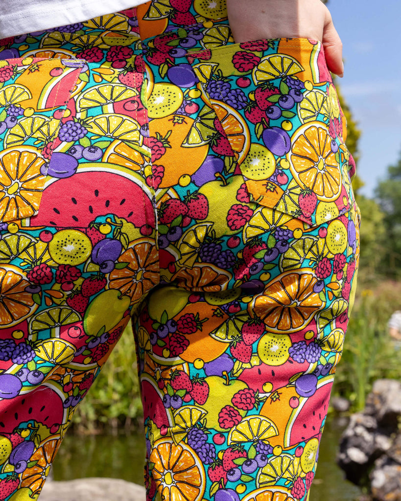 Close up of the Tutti Frutti Straight Leg High Waisted Jeans worn by a femme model stood outside on a rock pathway dotted over a pond with green foliage and flowers surrounding. She is facing forward with one hand in the pocket highlighting the front pockets, zip fly and run and fly button close. The 90s style trousers feature various brightly coloured fruit slices on a turquoise blue base.