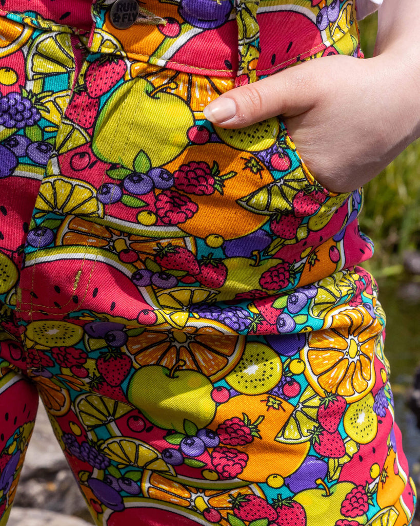 Close up of the Tutti Frutti Straight Leg High Waisted Jeans worn by a femme model stood outside on a rock pathway dotted over a pond with green foliage and flowers surrounding. She is facing forward with one hand in the pocket highlighting the front pockets, zip fly and run and fly button close. The 90s style trousers feature various brightly coloured fruit slices on a turquoise blue base.