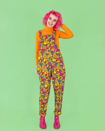 Lottie, a beautiful model with bright pink hair is wearing Run & Fly Tutti Frutti Stretch Twill Dungarees. The dungarees have a Jen James design of various fruit all over in bright red, orange, purple, yellow and green. The dungarees are paired with a long sleeved orange turtleneck top and pink boots. Lottie is smiling at the camera with one hand in the dungaree pocket and one hand in her hair. The background of the photo is lime green. 