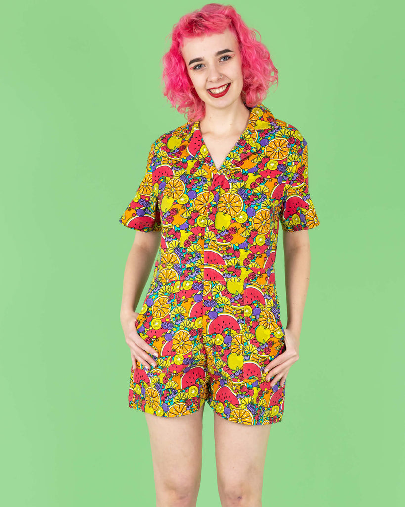 The Tutti Frutti Stretch Twill Playsuit on a femme model with short pink curly hair with pink jelly shoes and black socks. She is stood on a green background facing forward smiling with both hands resting in the pockets. The playsuit print is a blue background full of bright colourful fruit illustrations of limes, lemons, oranges, apples, berries and watermelon.