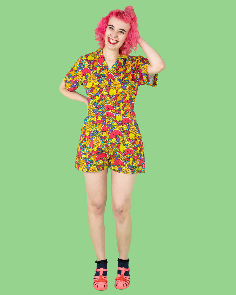 The Tutti Frutti Stretch Twill Playsuit on a femme model with short pink curly hair with pink jelly shoes and black socks. She is stood on a green background facing forward laughing with one hand on her hip and the other pushing up her hair. The playsuit print is a blue background full of bright colourful fruit illustrations of limes, lemons, oranges, apples, berries and watermelon.
