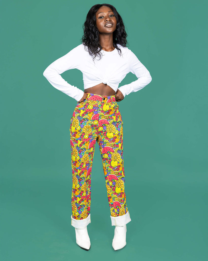 The Tutti Frutti Straight Leg High Waisted Jeans worn by a femme alternative style model with mid length black hair with a white long sleeve top and white cowboy boots on a teal studio background. She is facing forward smising to camera with both hands resting on her hips. The 90s style trousers feature various brightly coloured fruit slices on a turquoise blue base.