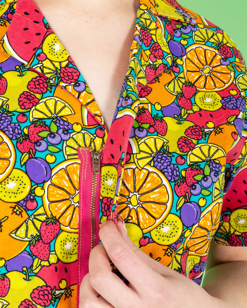 Close up of the Tutti Frutti Stretch Twill Playsuit on a femme model with short pink curly hair with pink jelly shoes and black socks. She is stood on a green background facing forward to highlight the zip up front detailing. The playsuit print is a blue background full of bright colourful fruit illustrations of limes, lemons, oranges, apples, berries and watermelon.