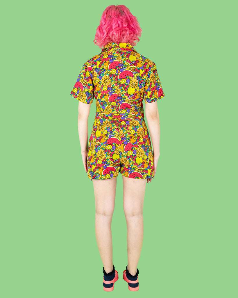 The Tutti Frutti Stretch Twill Playsuit on a femme model with short pink curly hair with pink jelly shoes and black socks. She is stood on a green background facing away with both hands resting by her side. The playsuit print is a blue background full of bright colourful fruit illustrations of limes, lemons, oranges, apples, berries and watermelon.
