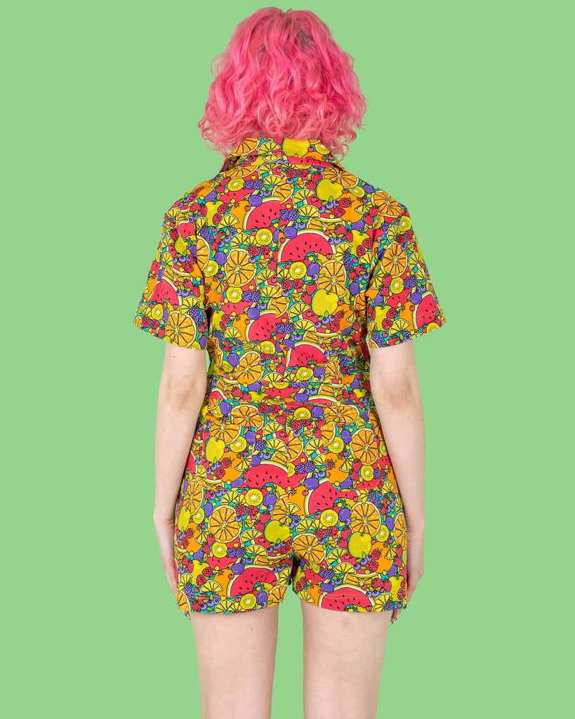 The Tutti Frutti Stretch Twill Playsuit on a femme model with short pink curly hair with pink jelly shoes and black socks. She is stood on a green background facing away with both hands resting by her side. The playsuit print is a blue background full of bright colourful fruit illustrations of limes, lemons, oranges, apples, berries and watermelon.