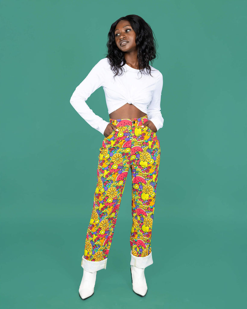 The Tutti Frutti Straight Leg High Waisted Jeans worn by a femme alternative style model with mid length black hair with a white long sleeve top and white cowboy boots on a teal studio background. She is facing forward smising to camera with both hands resting on her hips. The 90s style trousers feature various brightly coloured fruit slices on a turquoise blue base.
