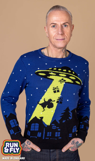 James is stood in front of award beige backdrop wearing the beam me up santa UFO jumper. He is facing forward smiling with both hands in his pockets. The jumper is a dark blue base with black house silhouettes running along the bottom and sleeve cuffs, a yellow and black ufo silhouette beaming up santa and presents with white snow dots. The back is plain blue