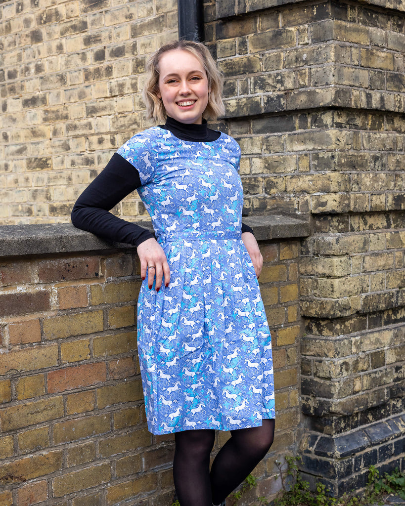 Femme model with short blonde hair is stood outside by a brick wall wearing Unicorn Tea Dress with a long sleeve black turtle neck underneath. The blue dress has an all over unicorn print. The model is stood leaning against a wall smiling at the camera with one leg bent. 