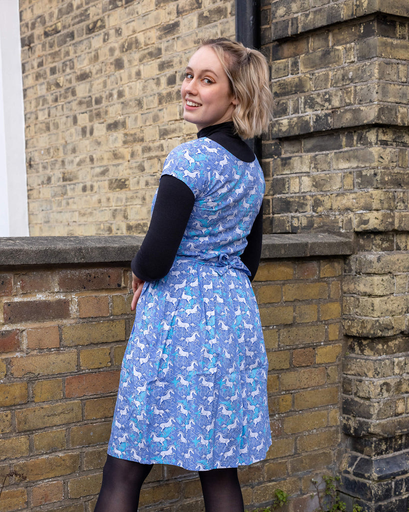 Femme model with short blonde hair is stood outside by a brick wall wearing Unicorn Tea Dress with a long sleeve black turtle neck underneath and black tights and boots. The blue dress has an all over unicorn print. The model is posing with her back to the camera and is looking back over her shoulder smiling at the camera.