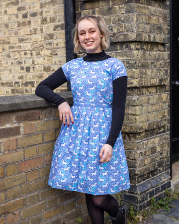 Femme model with short blonde hair is stood outside by a brick wall wearing Unicorn Tea Dress with a long sleeve black turtle neck underneath. The blue dress has an all over unicorn print. The model is stood leaning against a wall smiling at the camera with one leg bent. 