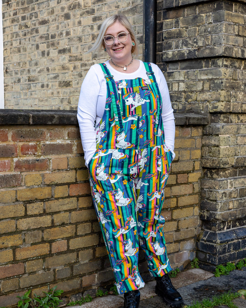 A person modelling Run & Fly unicorn dungarees, standing against a brick wall. The dungarees feature a fun print of colourful unicorns, stars, and rainbows on bold green and yellow stripes. Styled with black boots and a white long-sleeved top. Run & Fly focuses on ethical slow fashion, offering playful and eco-friendly clothing.