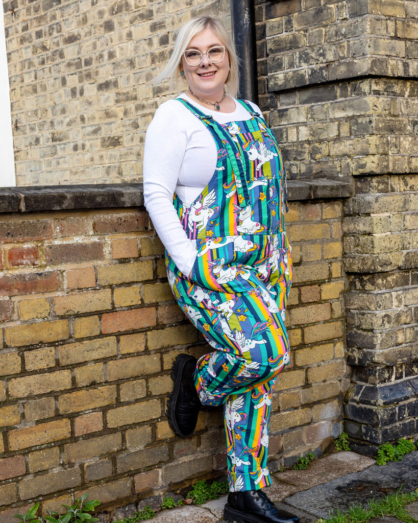 A smiling person wearing Run & Fly unicorn dungarees, standing against a brick wall. The dungarees feature a whimsical print of unicorns, stars, and rainbows on a green and yellow striped background. Paired with black boots and a white long-sleeved top. Run & Fly is a small ethical slow fashion company known for its quirky and sustainable designs.