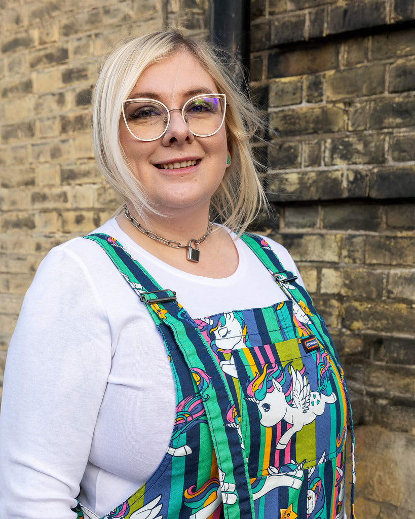 A close-up shot of a blonde hair femme model wearing Run & Fly unicorn dungarees, featuring vibrant rainbow-striped backgrounds and cartoon unicorns with rainbow manes. The model pairs the dungarees with a white long-sleeve top and silver accessories, including a lock necklace and bold glasses. Run & Fly is an ethical slow fashion brand, offering inclusive, quirky designs for all body shapes.