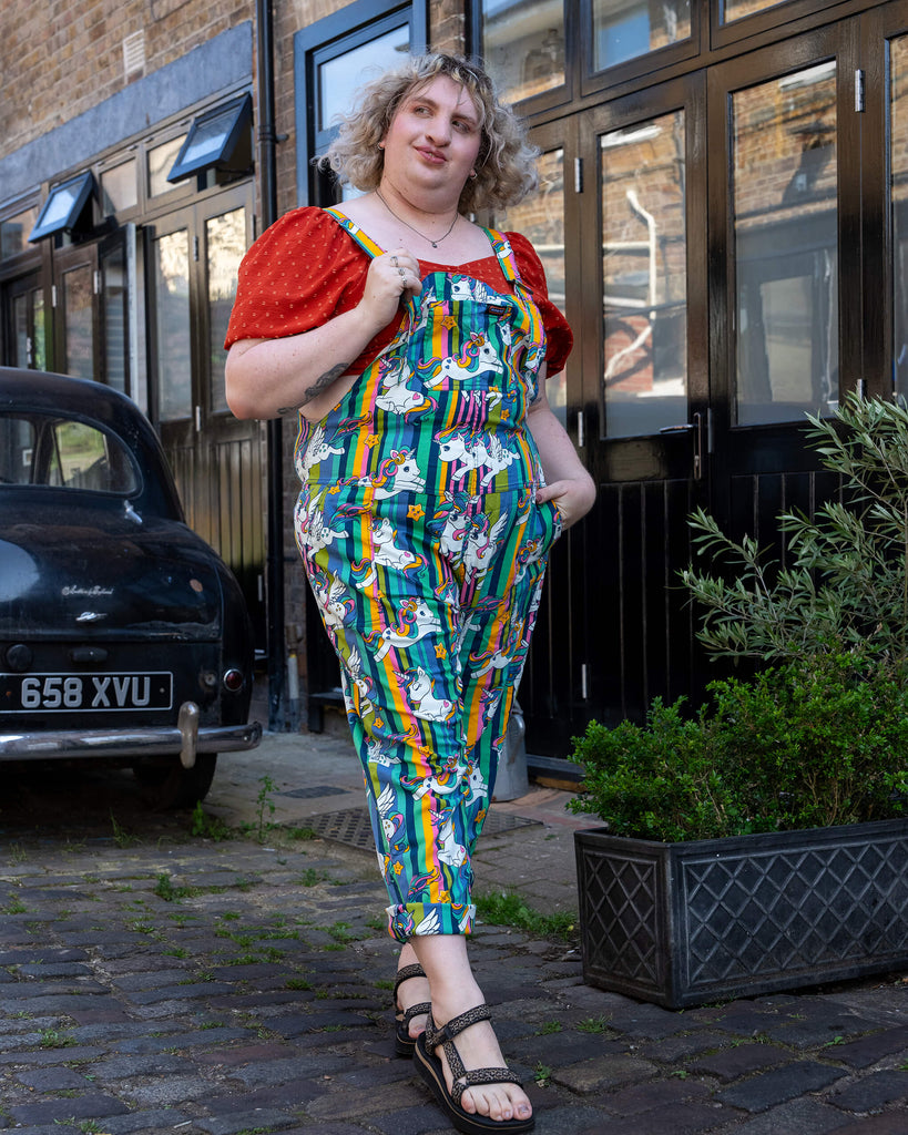A femme tall blonde haired model poses confidently in Run & Fly unicorn dungarees featuring a vibrant rainbow-striped background with cartoon unicorns and stars. Paired with a red puff sleeve top and black sandals, the outfit is playful and eye-catching. Run & Fly is a small ethical slow fashion brand known for quirky, colourful designs made to fit all body shapes and sizes.