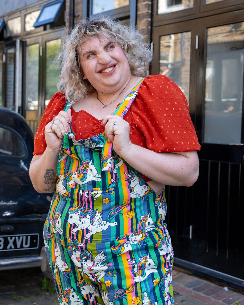 A smiling femme blonde haired model wearing Run & Fly dungarees with a bold unicorn print. The rainbow-striped dungarees feature fun cartoon-style unicorns with rainbow manes and stars. The model pairs the dungarees with a red puff sleeve top and adds fun accessories to complete the look. Run & Fly is a small ethical slow fashion brand, offering inclusive designs full of character and charm.
