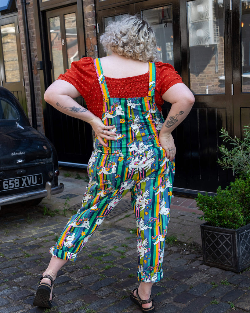 Back view of a femme short blonde haired model wearing Run & Fly unicorn print dungarees. The dungarees feature a rainbow-striped background with cartoon unicorns and stars. The model pairs the dungarees with a red polka dot puff sleeve top. Run & Fly focuses on ethical slow fashion with a quirky, colourful aesthetic, creating unique, playful clothing for all body types.