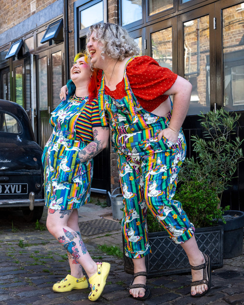 Two models laughing together while wearing Run & Fly unicorn print clothing. The femme model on the left has rainbow hair wearing a vibrant pinafore dress, and the femme model on the right has short curly blonde is styled in rolled-up dungarees with sandals. The rainbow-striped print is decorated with cartoon unicorns and stars. Run & Fly is a small, ethical slow fashion company known for its bold and inclusive designs.
